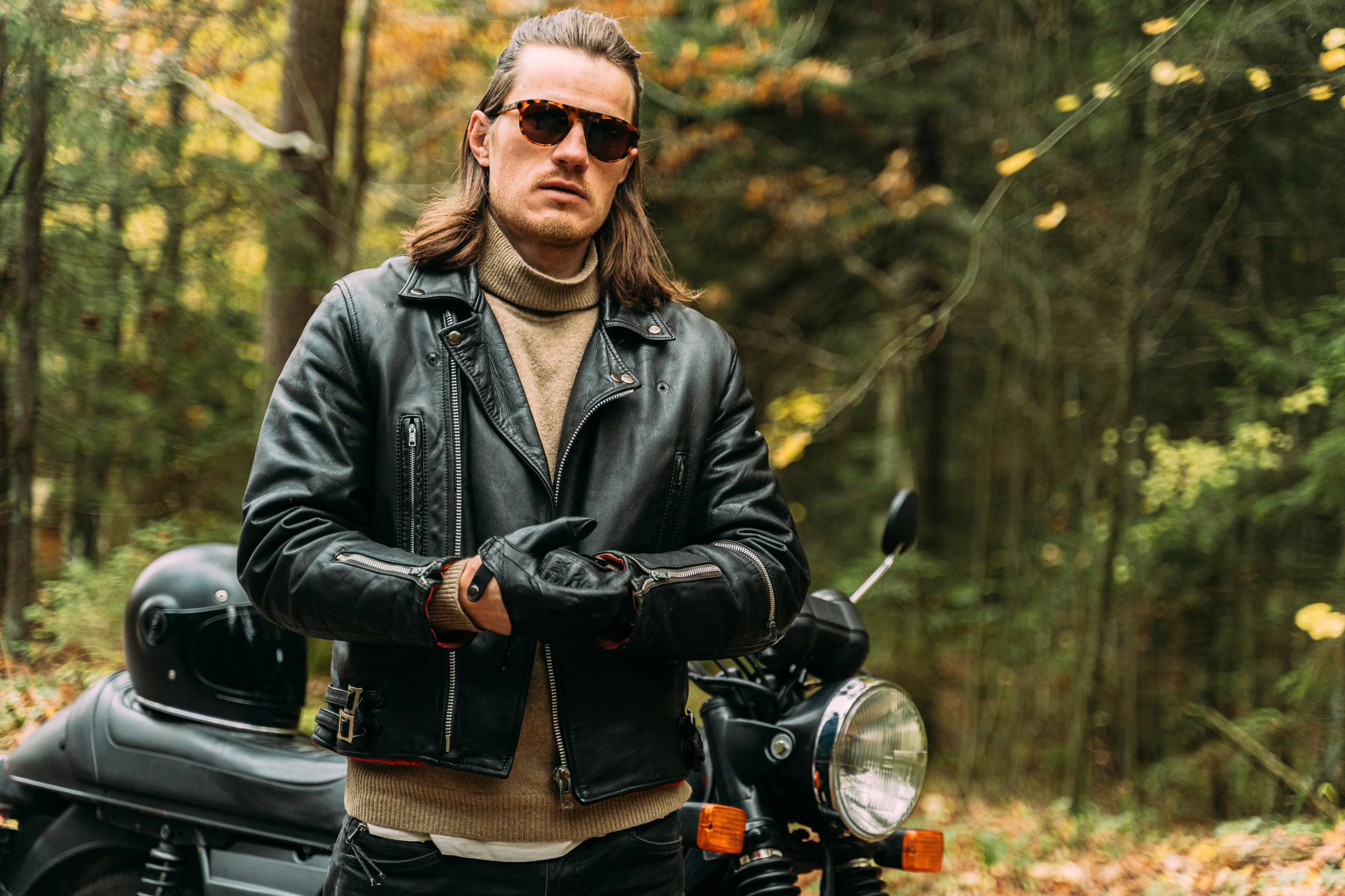 Man in a leather jacket standing next to a motorcycle in a forest setting